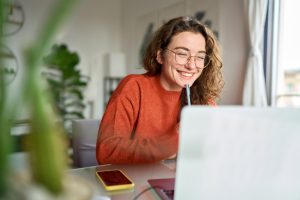 Junge Frau sitzt in einem Wohnraum lächelnd vor einem Laptop